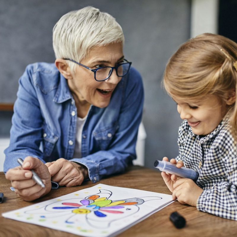 Grandmother and granddaughter sitting at table, painting colouring book Grandmother and granddaughter sitting at table, painting colouring book